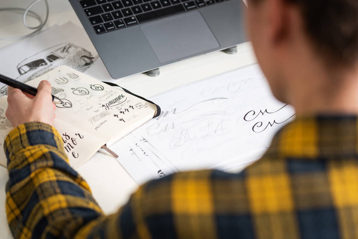 Graphic designer hand-sketching a bespoke corporate logo concept in a sketchbook at a Stockport creative agency, mapping out brand identity typography for a local business marketing campaign.